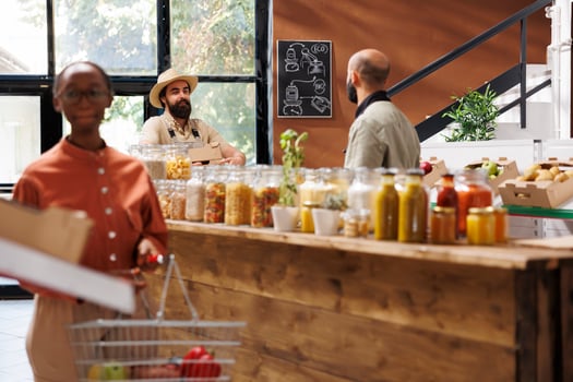 Black woman looks around in store while holding basket filled with food products. Focus on background are two men, one a farmer with boxes and another a store worker, interacting with each other. photo