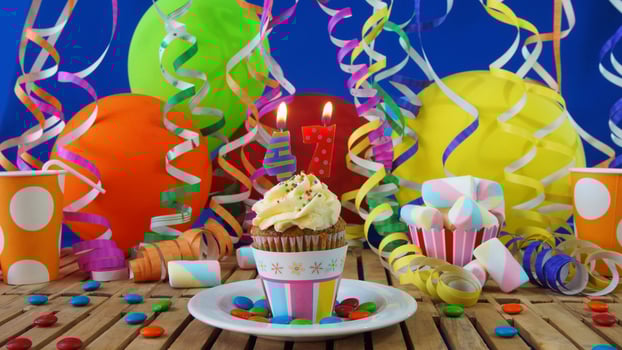 Birthday 47 cupcake with candles burning on rustic wooden table with background of colorful balloons, plastic cups and candies with blue wall in the background photo
