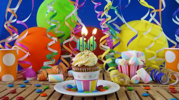 Birthday 66 cupcake with candles burning on rustic wooden table with background of colorful balloons, plastic cups and candies with blue wall in the background photo