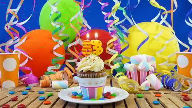 Birthday 83 cupcake with candles burning on rustic wooden table with background of colorful balloons, plastic cups and candies with blue wall in the background photo