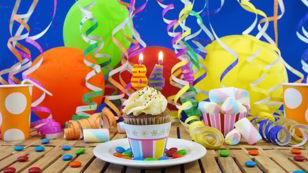 Birthday 84 cupcake with candles burning on rustic wooden table with background of colorful balloons, plastic cups and candies with blue wall in the background photo