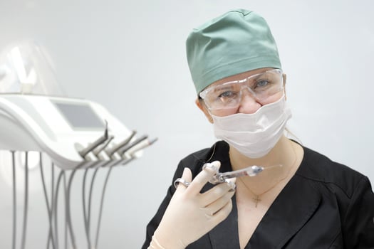 female doctor in a dental office in a mask and goggles Green cap to contain hair black suit in her hands carpal syringe and hygienic surgical gloves white wall latest technology cross on the chest photo