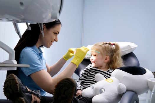 close-up face of a little girl and a doctor in a mask yellow hygienic gloves dentist tools pediatric dentistry sunglasses with teddy bear eyes girl treats teeth shows doctor painless treatment photo