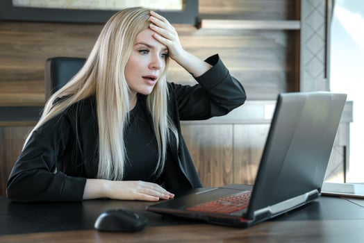 Woman with frightened expression looks at laptop screen, clutching her forehead with her hand in surprise photo