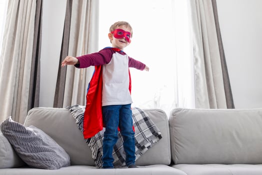 Boy in superhero costume with red cape and mask standing on couch. Indoor leisure and play concept photo
