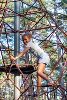 Joyful girl climbs a rope structure at a park, her smile expressing the thrill of adventure amidst tall pines, perfect for themes of active childhood, and sports activities. photo