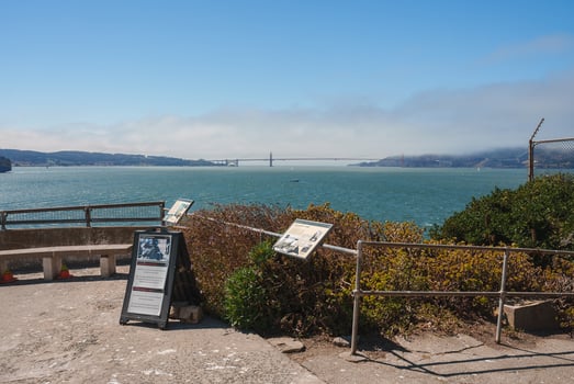 Scenic viewpoint from Alcatraz Island overlooking San Francisco Bay. Metal railing, informational signs, Golden Gate Bridge in misty background, lush vegetation. photo
