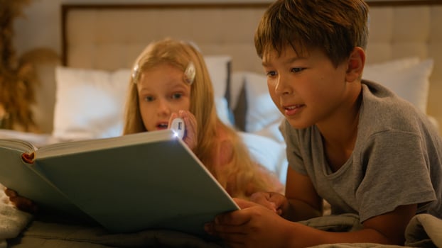 Caucasian children kids boy girl older brother younger sister lying on bed at home evening reading interesting book using flashlight read fairytale story for night bedtime happy family relationships photo