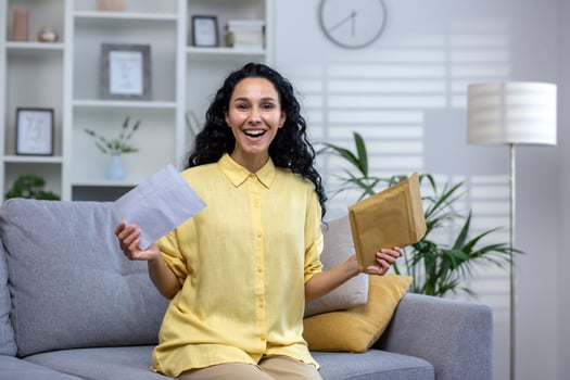 Happy curly-haired hispanic woman sitting at home on sofa in living room, holding open envelope. Cute brunette is happy with the news, winning lottery, happy news, smiling and looking at the camera. photo