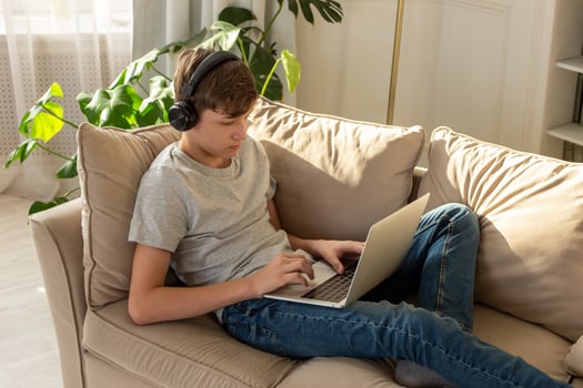 A teenage boy , lying on the couch in a sunny room, wearing black headphones, playing on a laptop photo