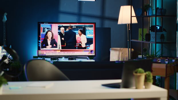 Apartment illuminated by RGB lights with TV broadcast showing Black Friday news story. Blurry foreground of work desk with laptop and mini house plants in dimly lit empty home studio interior photo