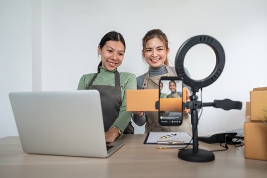 Two asian woman business owner at store showing parcel box in front of smartphone to recording vlog live streaming for sale online products to customer, Online shopping concept photo