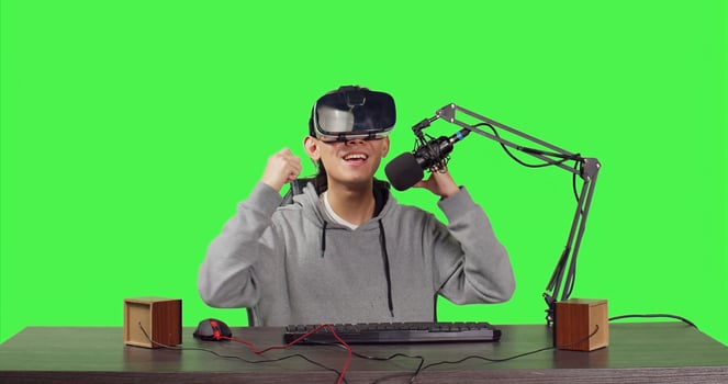 Streamer broadcasting live online games from his desktop while participating online using virtual reality equipment. Asian person streaming videogames, content creator on greenscreen. photo