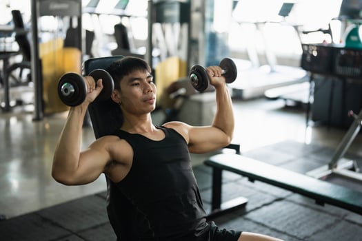 A focused man in a black tank top performs a seated dumbbell shoulder press in a modern gym. His muscular build and concentrated expression highlight his dedication to strength training. photo