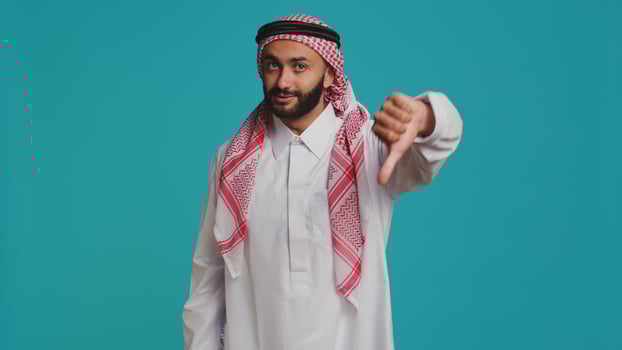 Islamic adult gives thumbs down and feeling disappointed about something, indicating muslim culture with traditional robe and scarf. Young person presenting rejection and negative sign. photo