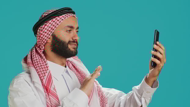 Arabic person engages in online conversation on his mobile device, wearing national costume. Young man with muslim headdress and thobe discussing on videoconference call in studio. photo