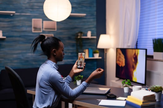 Woman with dark skin holding phone in online meeting sitting at desk in hime office late at night. Busy employee using modern technology network wireless doing overtime for job. photo