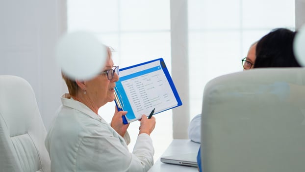 Elderly physician is brainstorming to discuss medical issues pointing on clipboard presenting list of sick patients. Team of doctors working in hospital office discussing symptoms of disease photo