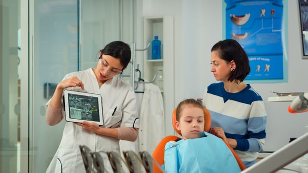 Pediatric dentist showing teeth x-ray on tablet pc computer screen to mother of patient girl at dental clinic office. Orthodontist using tablet explaining dental digital radiography to woman photo