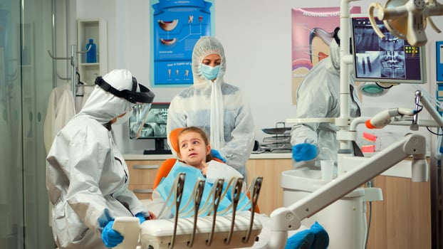 Stomatological team in protection suit preparing dental sterilized tools for children examination, mother standing near dentist chair. Concept of new normal dentist visit during coronavirus outbreak photo