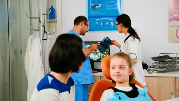 Team of stomatologist doctors analysing teeth x-ray of little patient with toothache sitting on stomatological chair talking with her mother. Doctor discussing with nurse about kid's dental health. photo