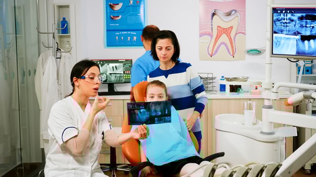 Stomatologist explaining dental treatment holding radiography pointing on affected teeth while man assistant preparing sterilized tools for surgery. Doctor and nurse working in stomatological unit photo