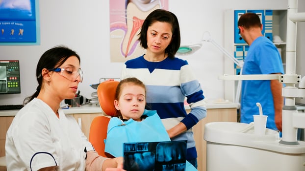 Pediatric dentist showing dental problems holding radiography pointing on affected teeth while man assistant preparing sterilized tools for surgery. Doctor and nurse working in stomatological unit photo