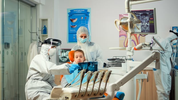 Pediatric dentist wearing protection suit treating girl patient in new normal stomatological unit showing teeth x-ray. Medical team wearing face shield coverall, mask, gloves, explaining radiography photo