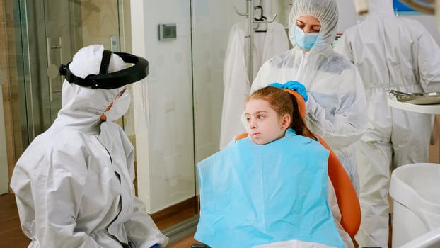 Little girl with dental bib and ppe suit listening pediatric dentist in coverall sitting in dental chair during coronavirus pandemic. Stomatologist wearing protection suit, face shield mask gloves photo