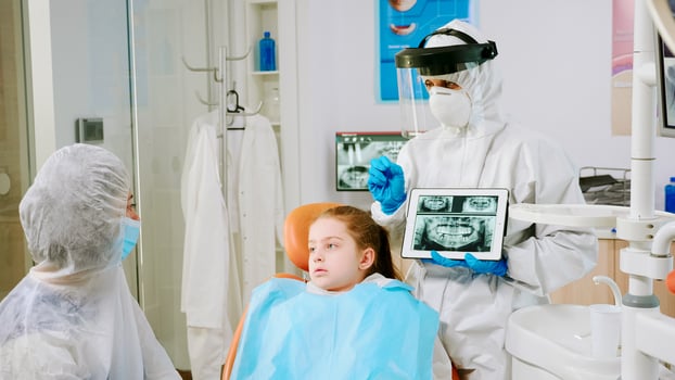 Dentist with face shield explaining panoramic mouth x-ray image to mother of kid patient during global pandemic. Stomatologist talking with woman wearing suit, coverall, protection suit, mask, gloves photo