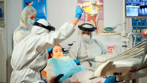 Dentist technician in coverall drilling kid tooth in dental clinic during coronavirus pandemic. Concept of new normal orthodontic visit in coronavirus outbreak wearing protective suit and face shield photo