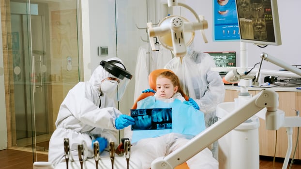 Dentist with coverall holding mouth x-ray image child patient talking with patient mother during global pandemic. Assistant and doctor talking wearing suit, coverall, protection suit, mask, gloves photo