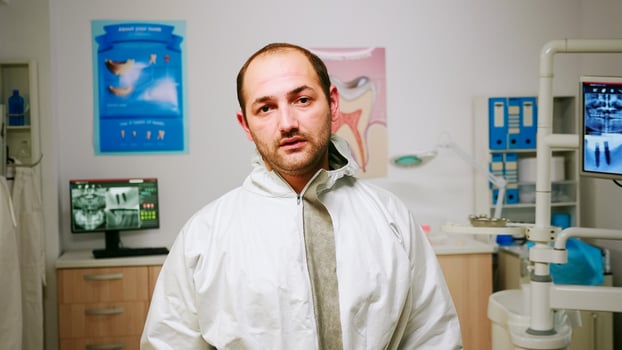 Portrait of exhausted stomatologist man taking off face shield looking at camera sitting in new normal dental office. pediatric nurse talking with child patient in background. photo