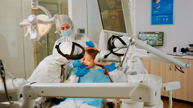 Child on dental chair in dentist surgery treated with dental drill during covid-19 pandemic. Orthodontist lighting the lamp wearing face shield, protection suit, mask and gloves talking with patient photo