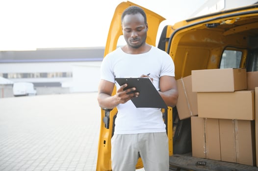 african american Worker unloading boxes from van outdoors. Moving service concept. Male employee of moving company makes inventory of packed boxes and writes data to clipboard. photo