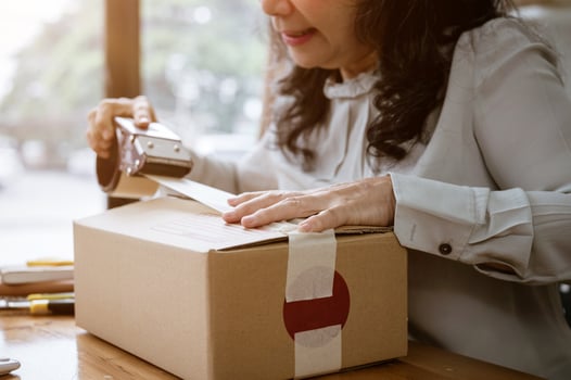 A mature Asian female online seller packing a shipping cardboard box, using a packing tape dispenser photo