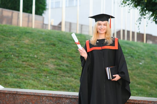 Happy cute caucasian grad girl is smiling. She is in a black mortar board, with red tassel, in gown, with nice brown curly hair, diploma in hand photo