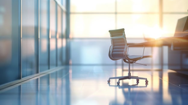 An empty swivel chair sits at a desk in a modern office with glass walls, bathed in warm sunset light. photo