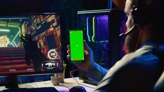 Man watching tutorial on green screen phone while playing first person shooter videogame with gun shooting laser bullets. Gamer learning to play game by looking at online guide on smartphone photo