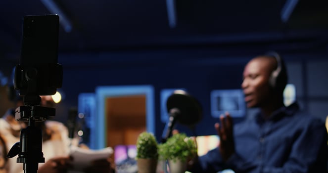 Focus on smartphone on tripod recording vlogger show host in blurry background interviewing guest in neon lights studio. Presenter records online podcast episode with mobile phone, close up photo
