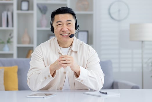 Cheerful Asian man wearing a headset and a casual beige shirt, enjoying a lively conversation while seated in a bright, modern living room. photo