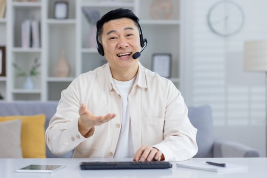 Cheerful Asian male in a beige shirt using a headset in a home office, looking at camera with a smile, possibly in a customer service call. photo