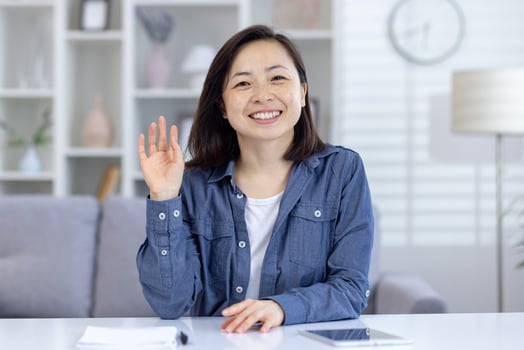 Smiling Asian woman sitting at her desk and waving at the camera. This image portrays a friendly conversation during a remote call Perfect of communication and modern workplaces at home. photo