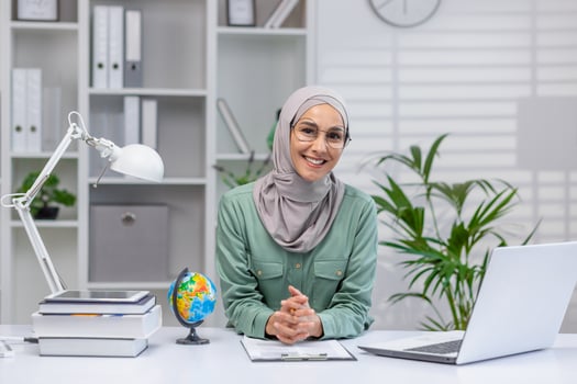 Portrait of happy teacher, woman in hijab sitting at desk smiling and looking at camera, muslim woman working remotely, teaching students and pupils using laptop in home office. photo