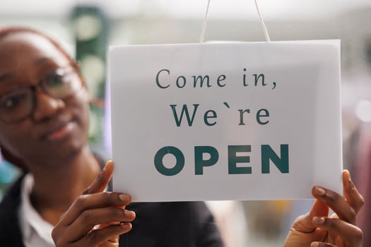 Fashion clothes shop manager holding opening signboard with come in message. Apparel store young african american woman assistant hanging sign informing clients about workday beginning photo
