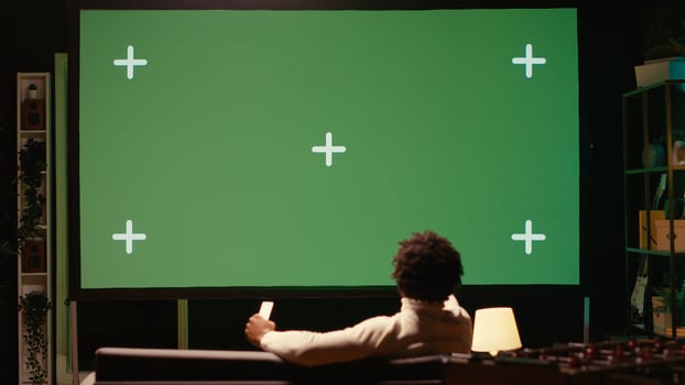 African american man using projector to binge TV series on subscription based streaming services. Cord cutter enjoying VOD shows on gigantic green screen television set photo