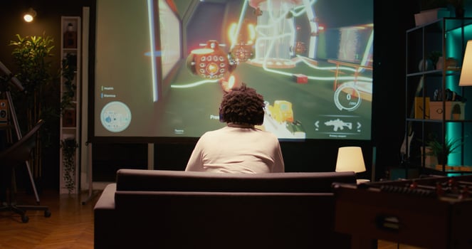 Man watching gameplay footage of videogame competition on gigantic smart TV, relaxing on couch. BIPOC gamer in home theatre following gaming tournament on television set, zoom out shot photo