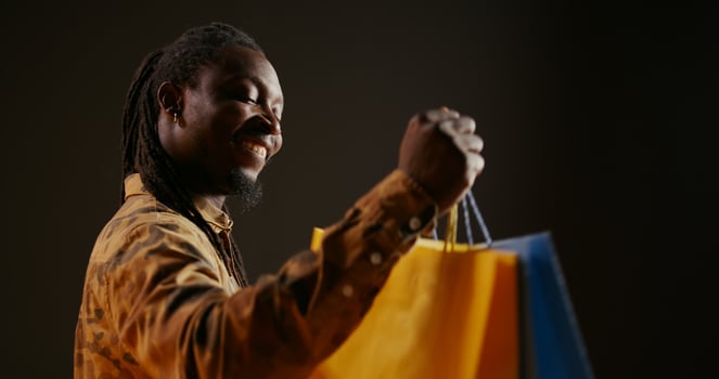 Happy pleased person carrying shopping bags after buying favorite clothes on discount, enjoying sales season at the mall. African american shopaholic guy looks at his new clothing items in bags. photo