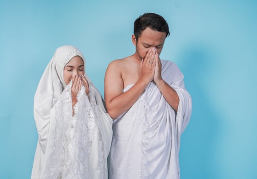 Young Asian Muslim couple wearing ihram and prayer gown praying with hand gesture over isolated by blue background. Hajj concept photo