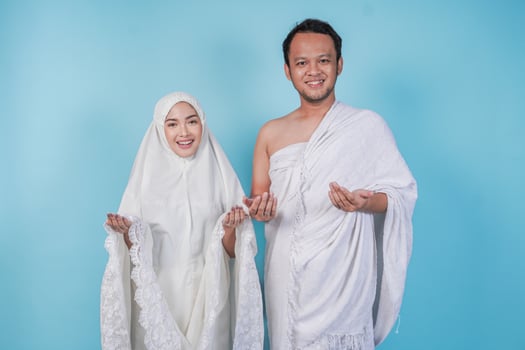 Smiling young Asian Muslim couple wearing ihram and prayer gown with praying hand gesture over isolated by blue background. Hajj concept. photo